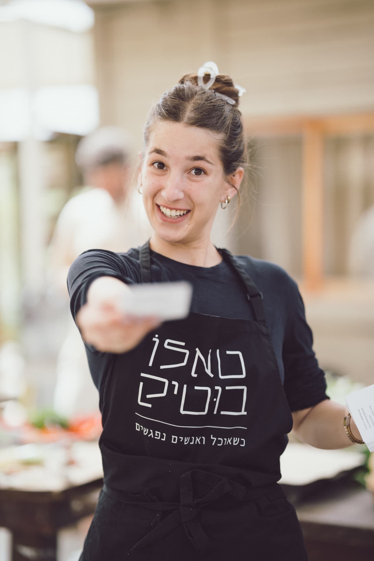 ליאורי מהצוות מחייכת ומושיטה כרטיס לעבר המצלמה כשהיא חוגרת סינר טאלו בוטיק