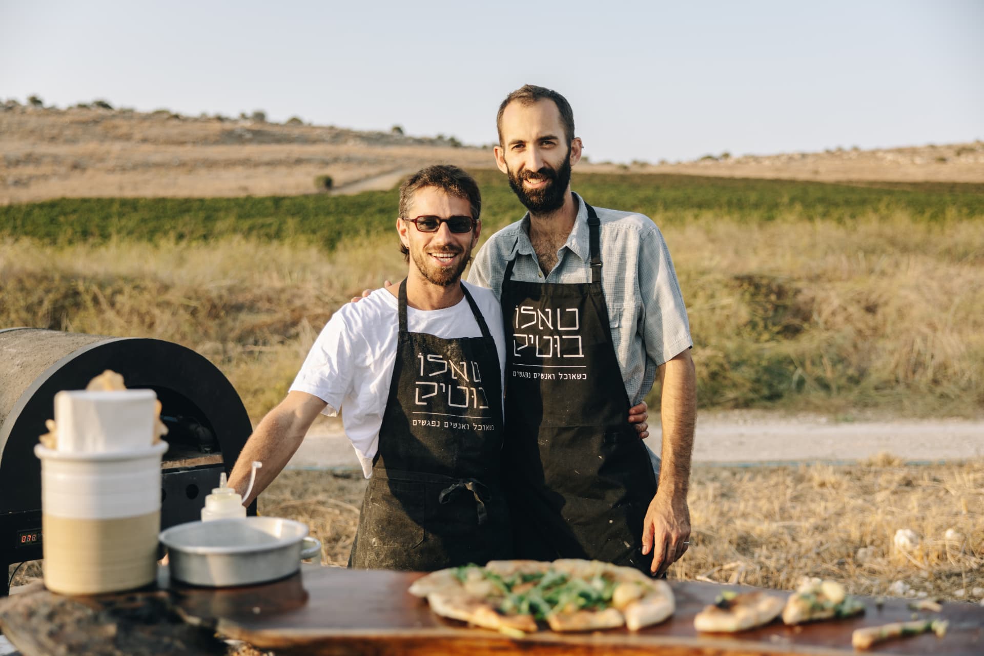 שני השפים עומדים זה לצד זה מאחורי דלפק עץ בשדה פתוח, חיוך ופיצה טרייה לפניהם