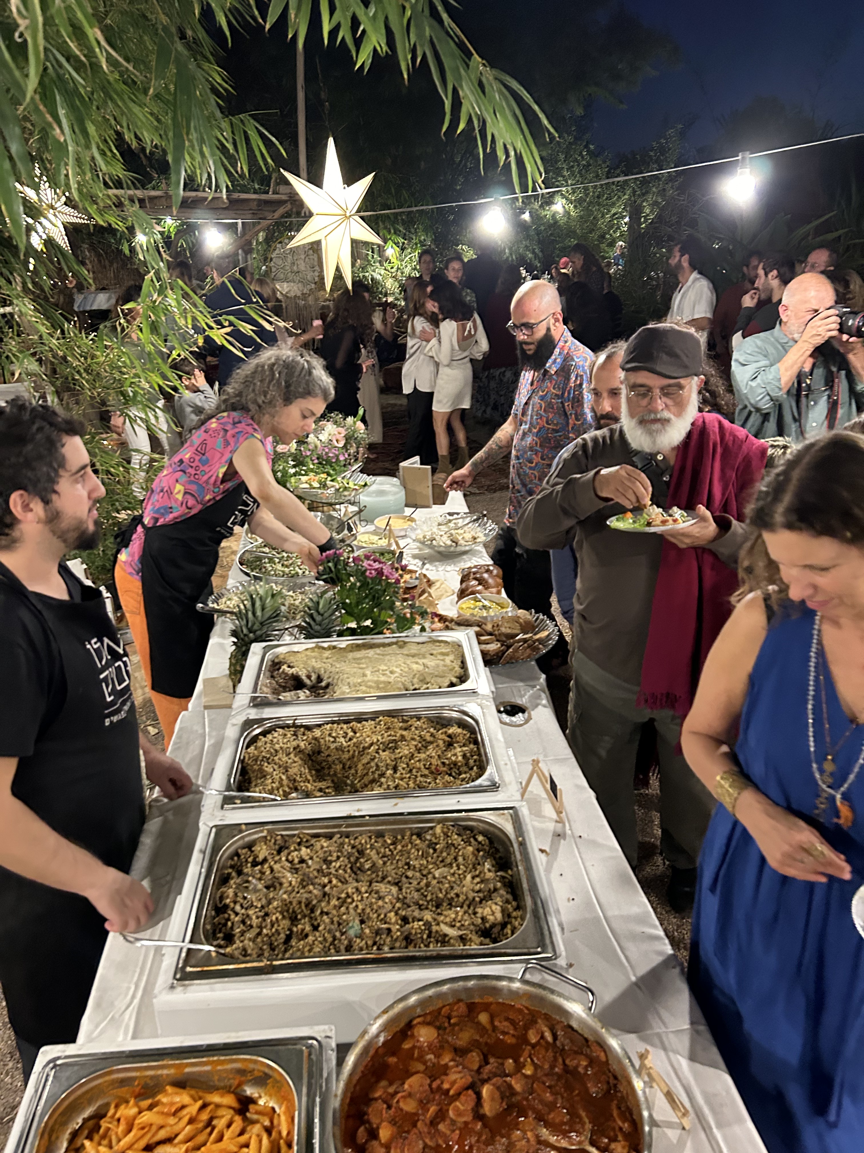 אורחים ניגשים לשולחן בופה בשעות הערב, עם צוות הגשה ומדורות תאורה
