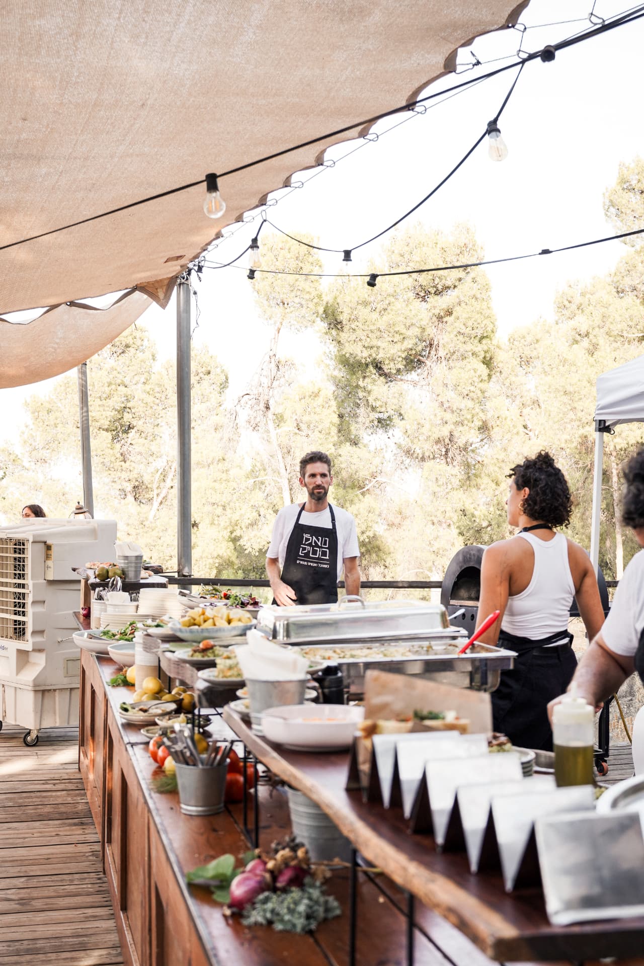 צוות ההגשה מאחורי שולחן המנות בחוץ, תחת קורת צל עם נורות
